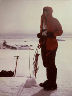 A woman in a red jacket stands atop a snow-covered mountain, holding climbing gear