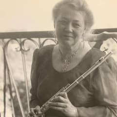 A black-and-white photo of a woman wearing an ornate necklace and her hair in a bun. She is gracefully holds a flute, showcasing her musical talent and love for the instrument.